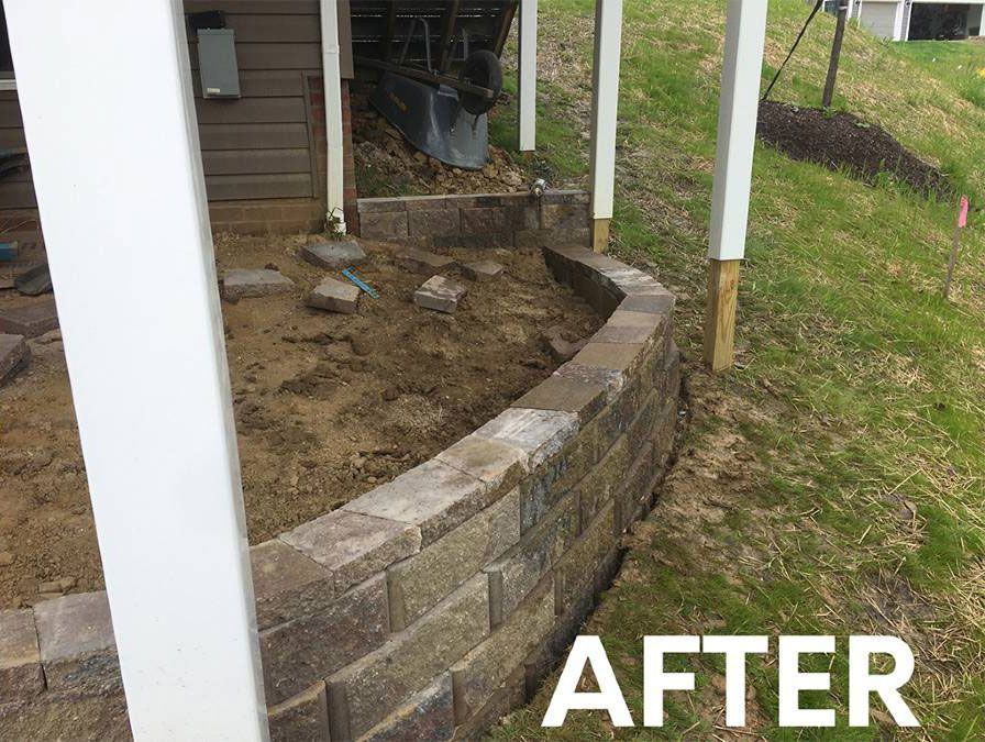 A low retaining wall made of tan and brown stone blocks built under a wooden deck in a grassy backyard.
