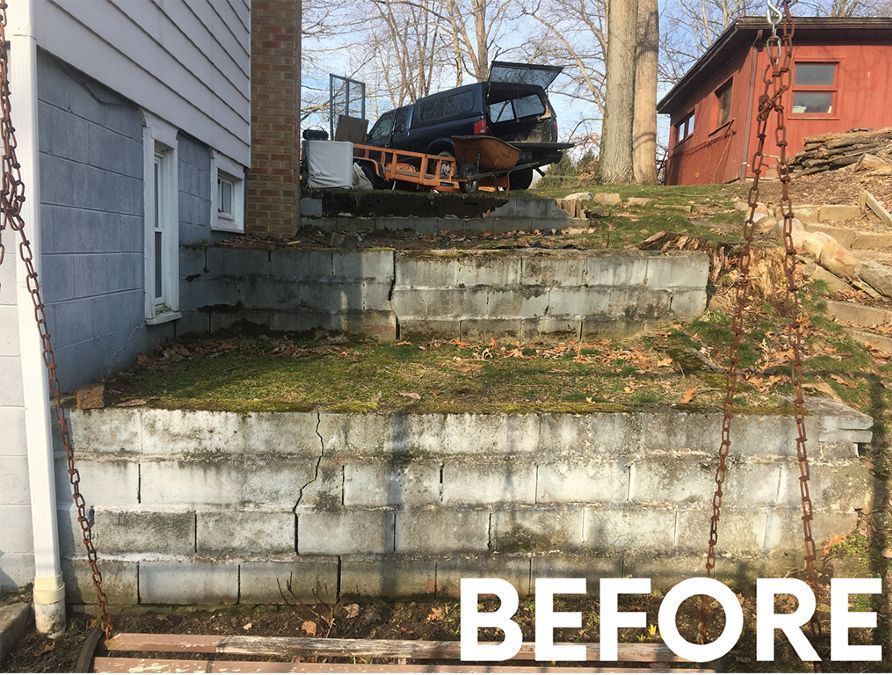 A tiered cinder block retaining wall next to a house, with a parked truck visible at the top level and a 