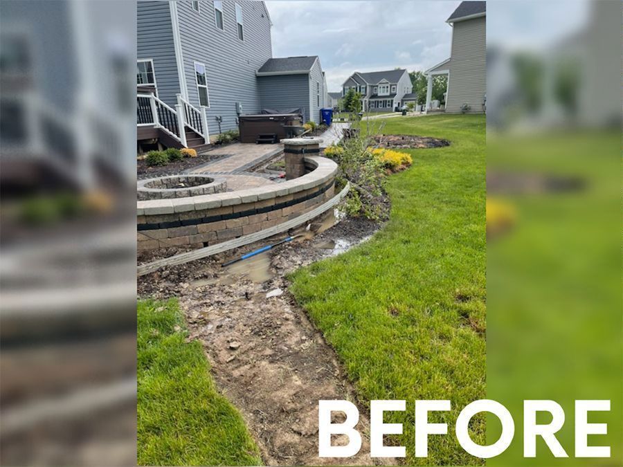 A backyard landscape under construction with stone retaining walls, a fire pit, and dirt pathways between patches of grass.