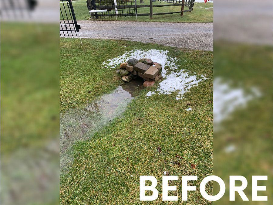 A stone-covered drainage pipe in a grassy yard during a snowy, wet day, marked with the word 