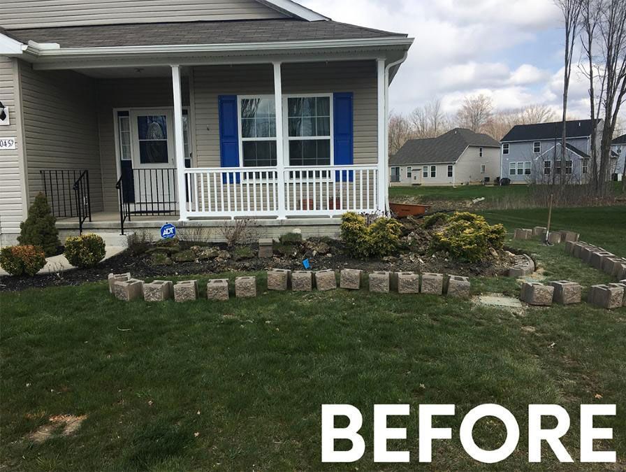 A beige house with blue shutters, a white porch, and a yard with square stone edging, labeled 
