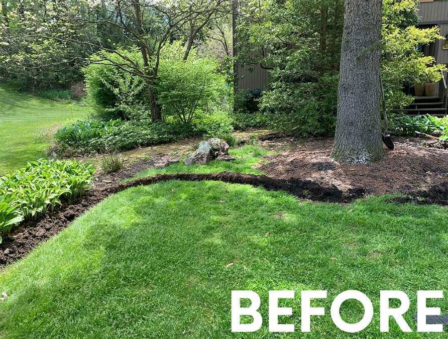 A grassy yard with a curved, freshly dug garden bed edge bordering a mulched area under trees, labeled 