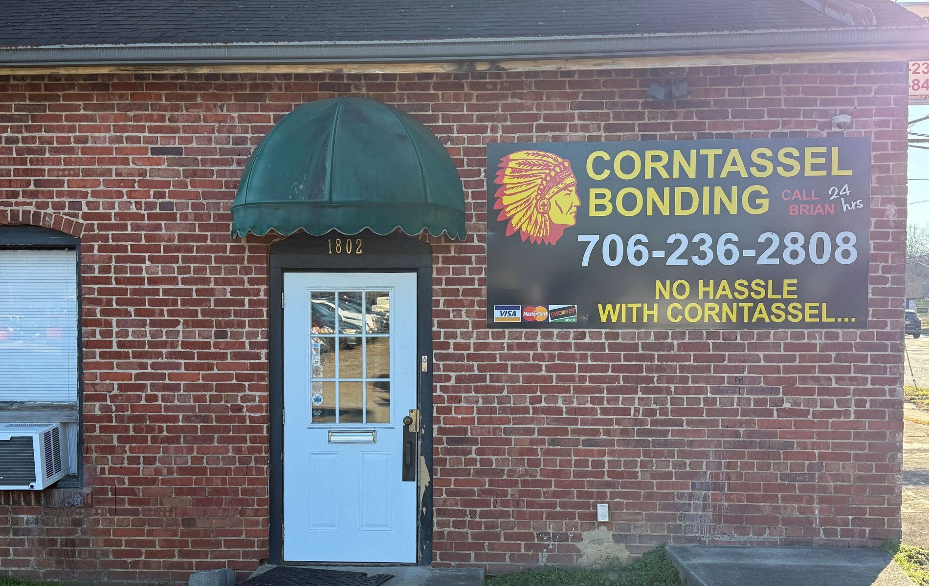 Brick building with green awning and sign for Corntassel Bonding. White door and air conditioner unit.