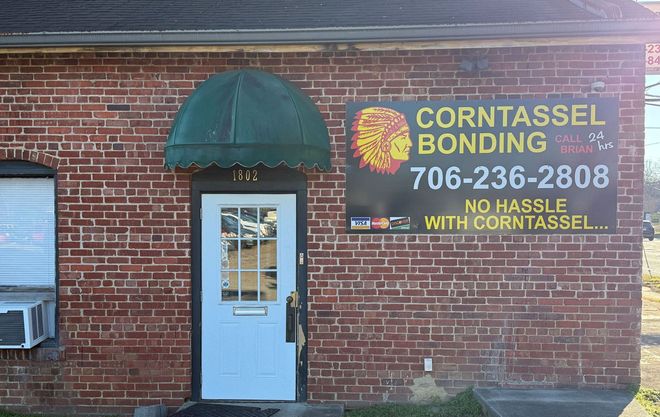 Brick building with green awning and sign for Corntassel Bonding. White door and air conditioner unit.