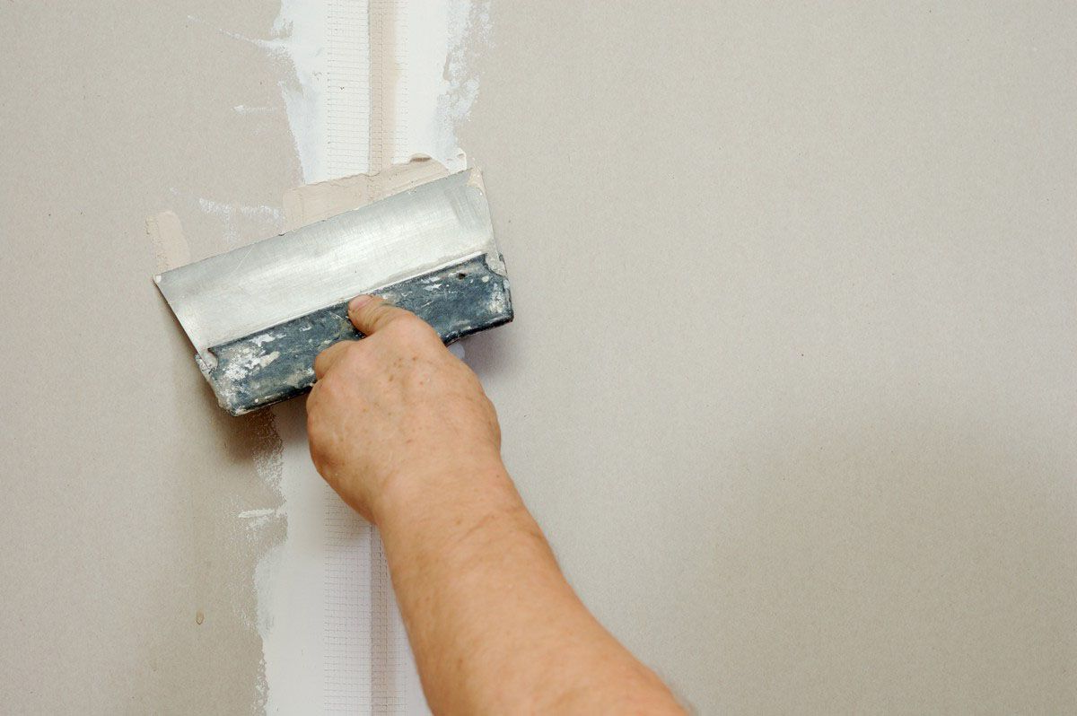 Hand smoothing white drywall compound on a beige wall with a putty knife