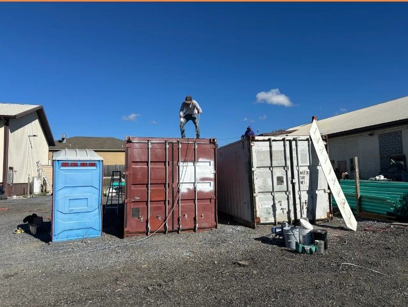 Person standing on top of a red shipping container in an industrial yard, with blue and white portable toilets nearby