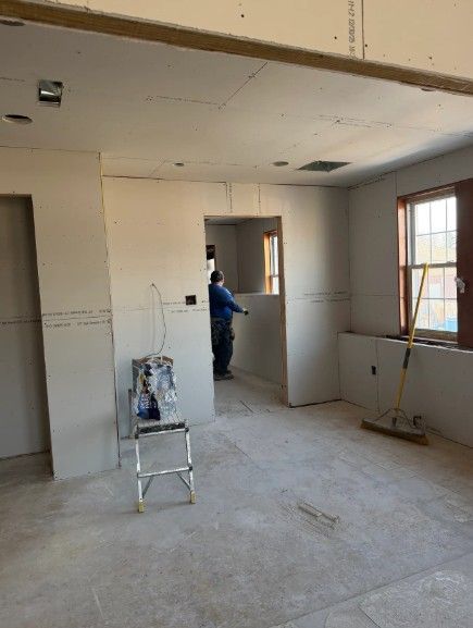 Unfinished room under renovation with drywall, ladders, and a worker near a doorway.