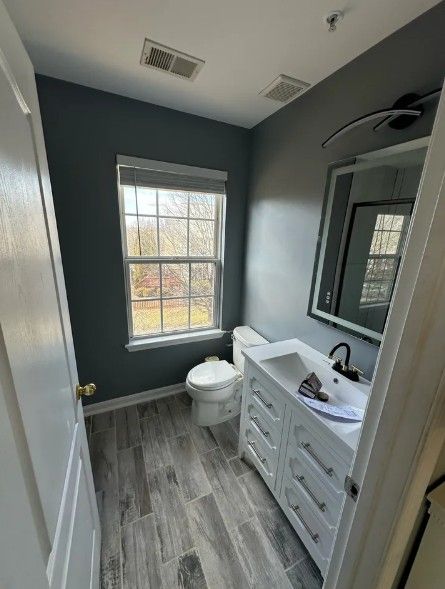 Small gray bathroom with window, toilet, vanity sink, and mirror.