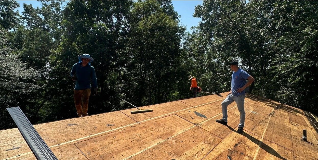 Workers installing plywood roof sheathing on a house under construction, surrounded by trees.