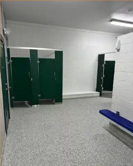 Public restroom with green stall doors, white tiled walls, and a blue bench on a speckled floor