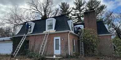Brick house with a steep roof and multiple ladders leaning against it under bare trees