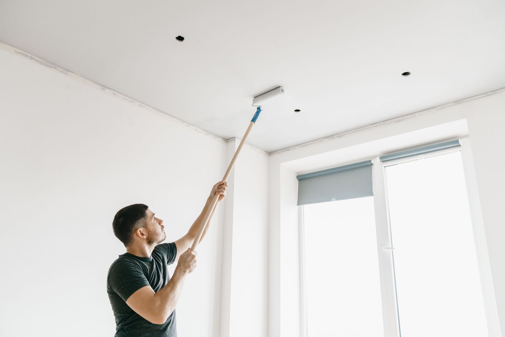 Person painting a white ceiling with a roller near a window in a bright room