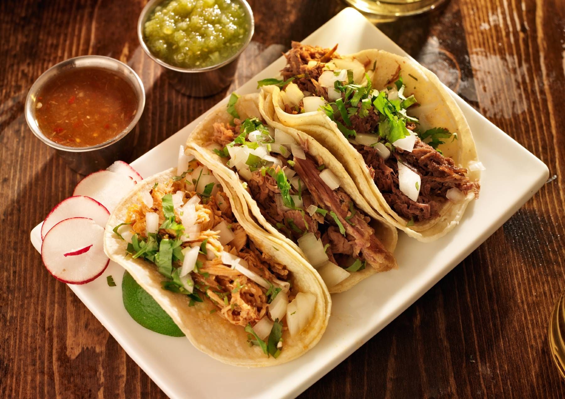 Three tacos are sitting on a white plate on a wooden table.