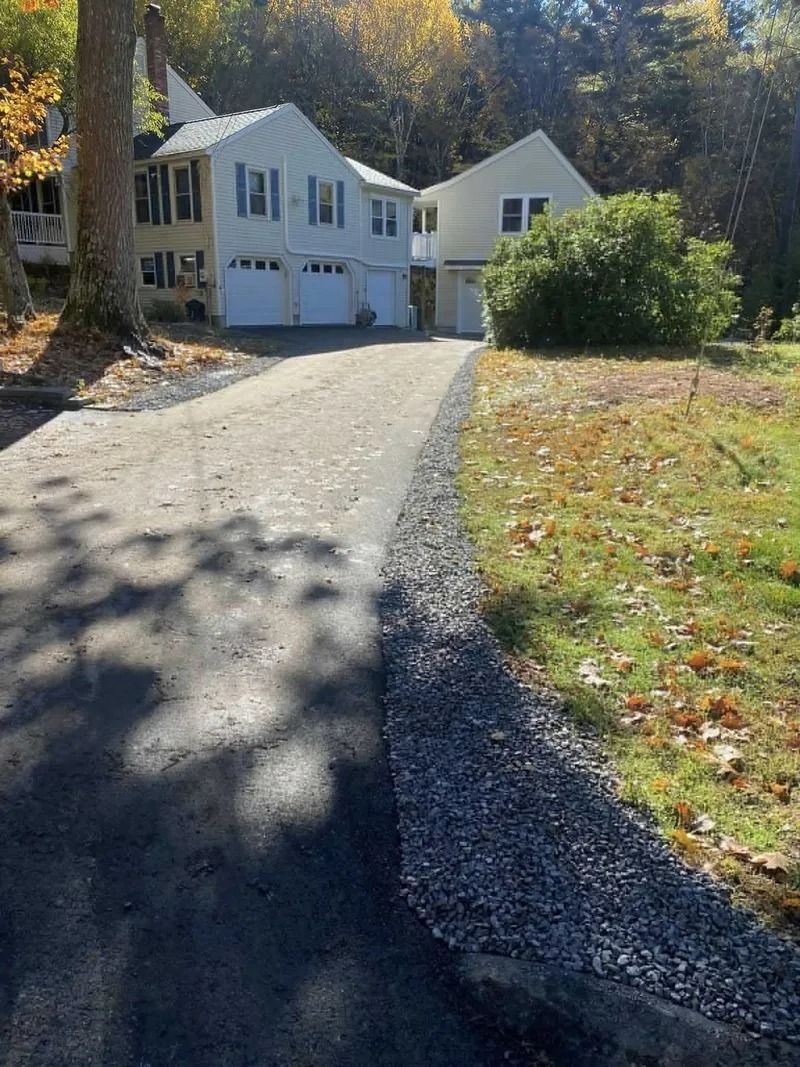 Asphalt driveway leading to a light-colored house with a two-car garage, in a yard with fall foliage.