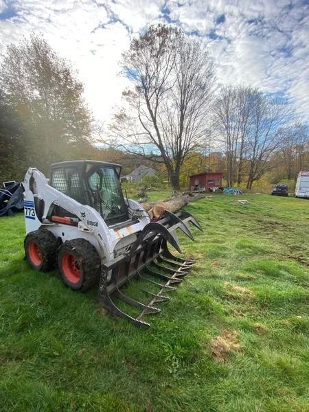 Bobcat skid-steer with grapple attachment on a grassy field, holding a log, trees and a building in the background.