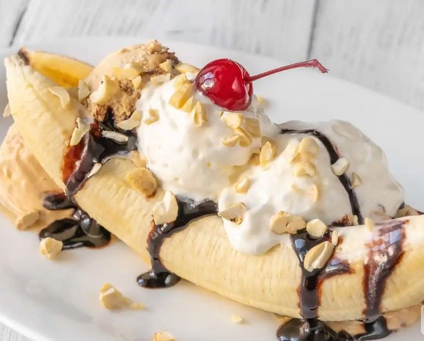 A banana split with three scoops of ice cream, chocolate sauce, chopped nuts, and a maraschino cherry on a white plate.