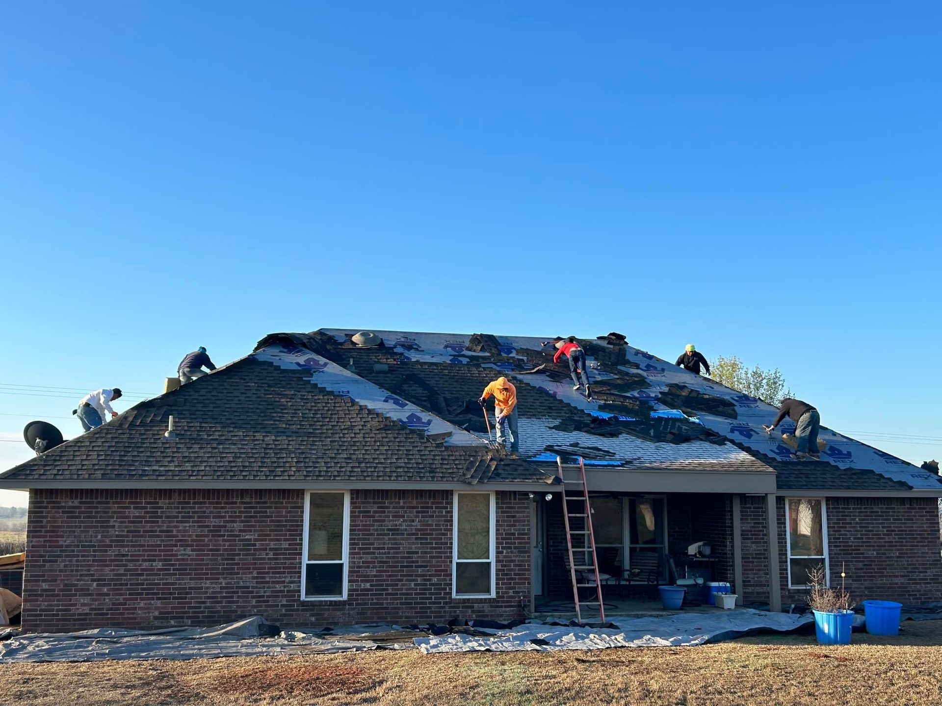 A crew works on replacing the roof of a single-story brick house under a clear blue sky.