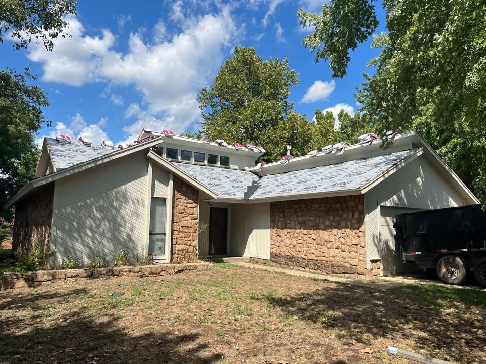A house with stone siding under a partially shingled roof, with a dumpster trailer parked in the driveway.