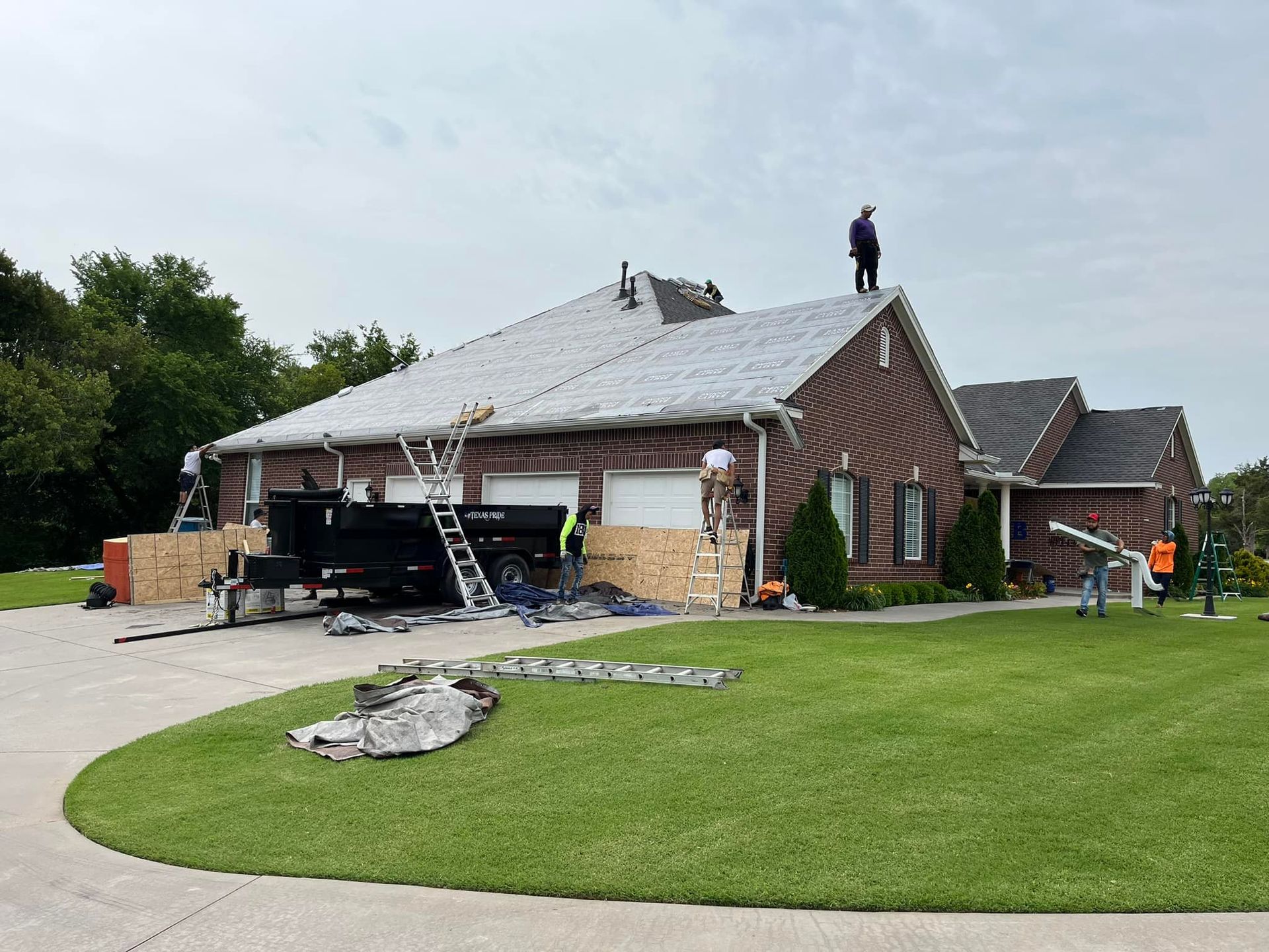 Workers perform roof repairs on a brick house, using a ladder and a dumpster trailer in a residential yard.
