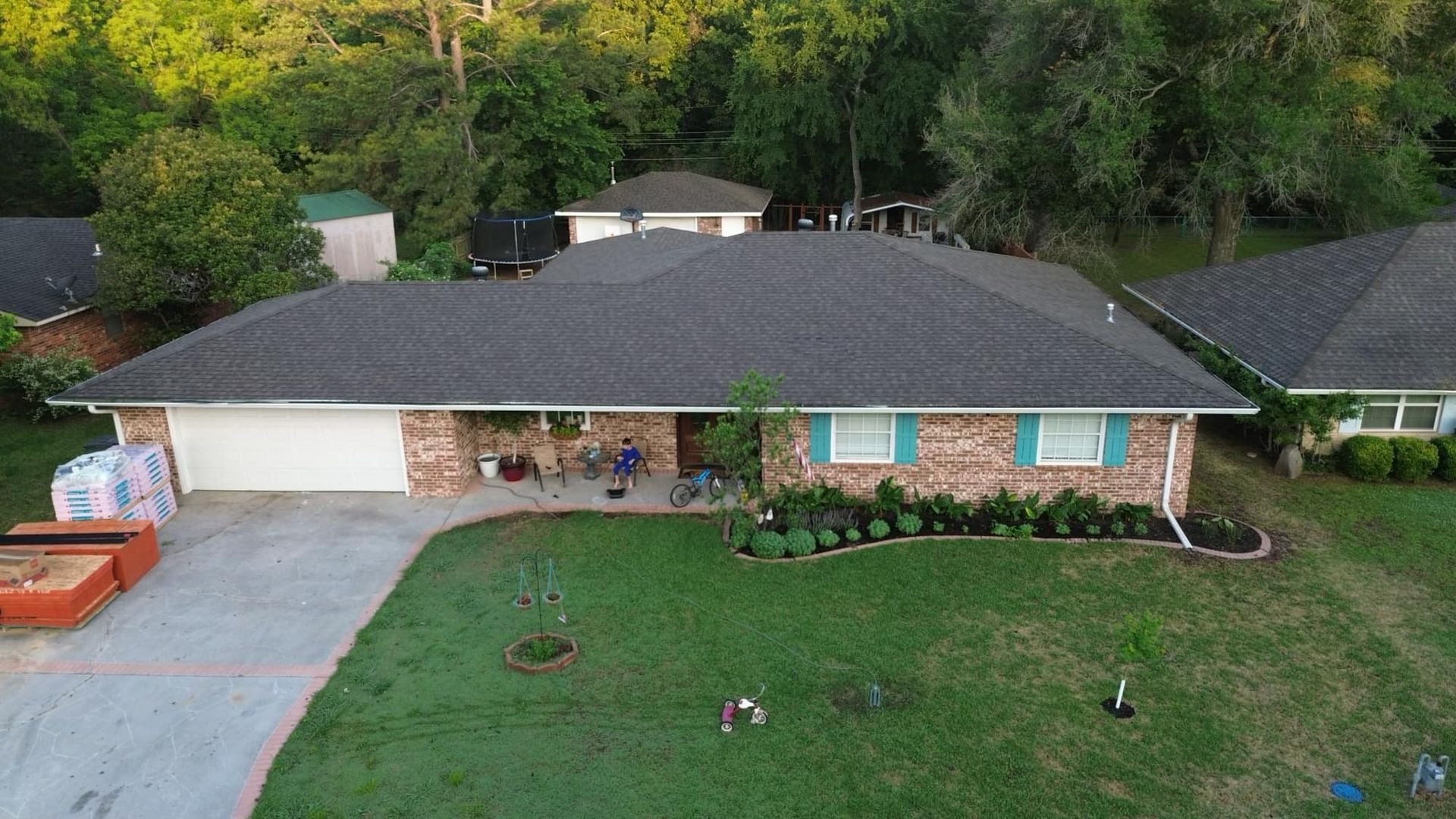 Aerial view of a single-story brick house with a dark shingled roof, teal shutters, and a lawn in a residential area.