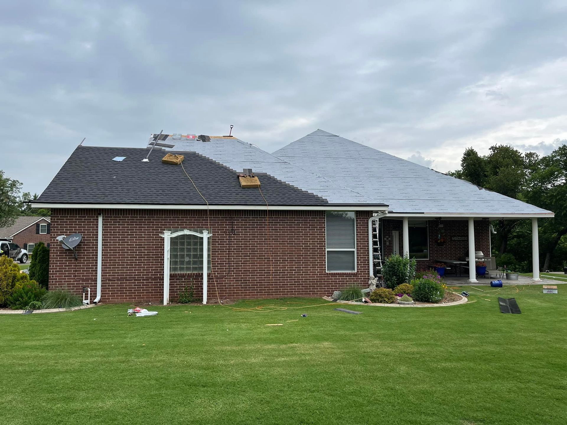 A brick house with a partially shingled roof under repair, showing exposed underlayment on a cloudy day.