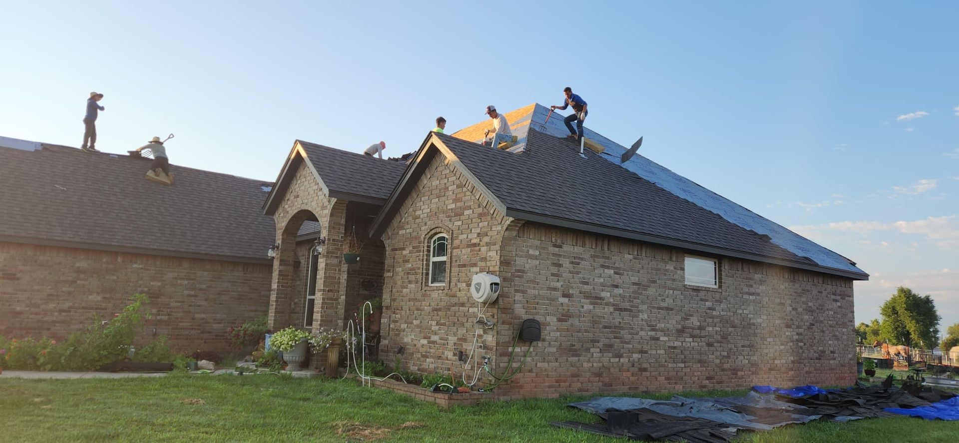 A group of workers are installing new shingles on the roof of a stone-faced house on a sunny day.