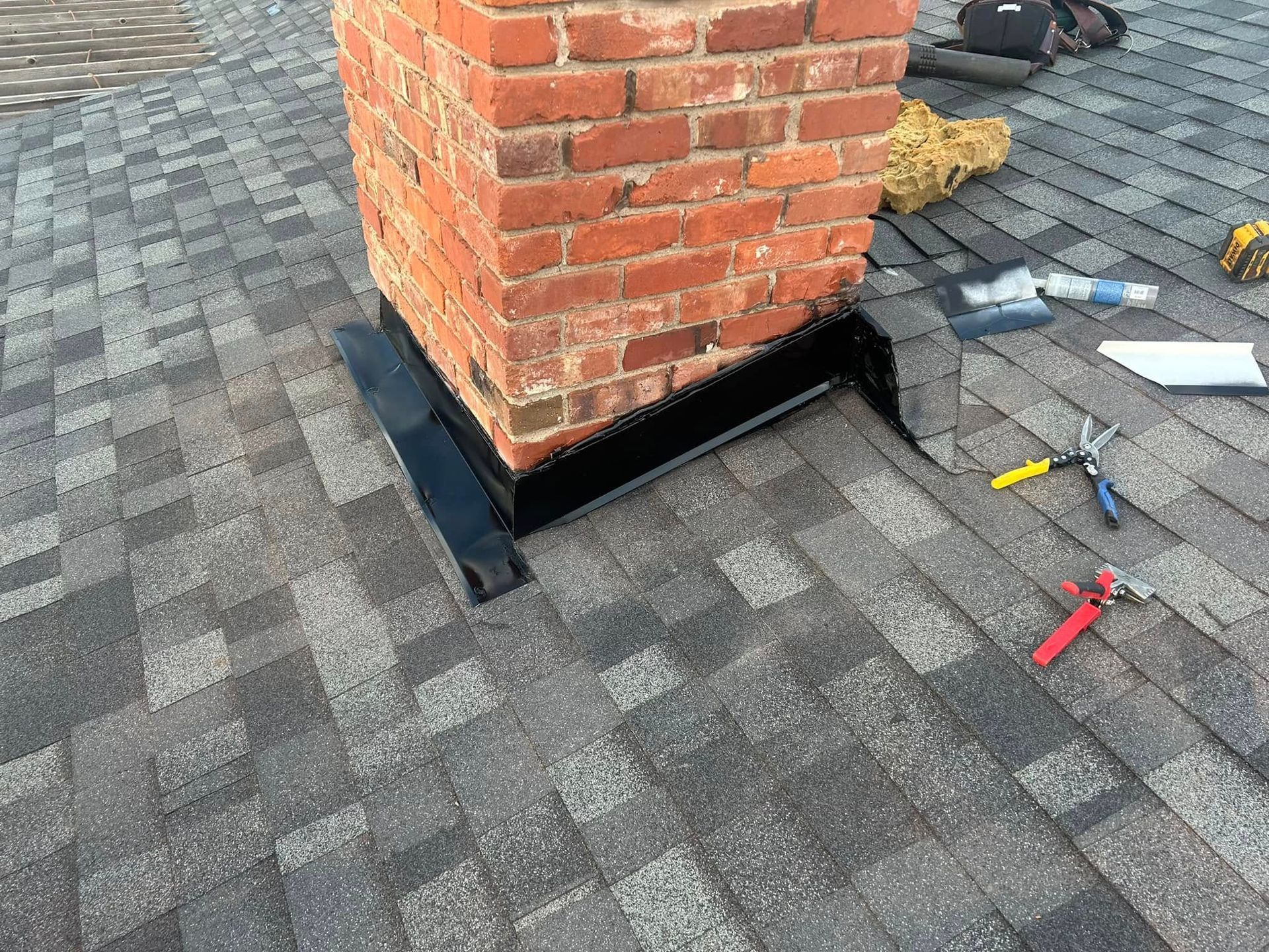 Black flashing installed around a brick chimney on a shingled roof, with various construction tools nearby.