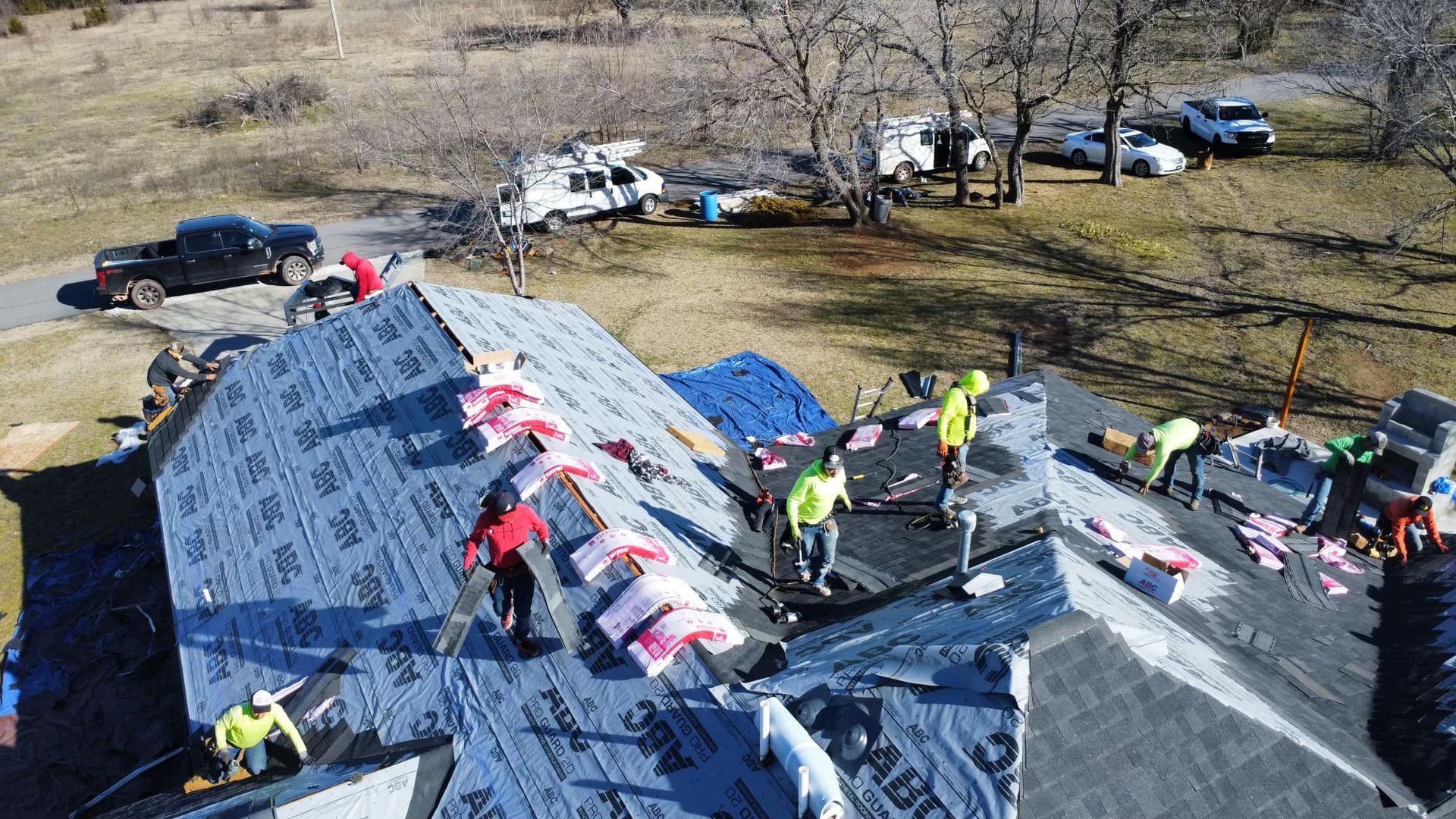 Aerial view of workers in neon shirts installing underlayment on a residential roof during a sunny day.
