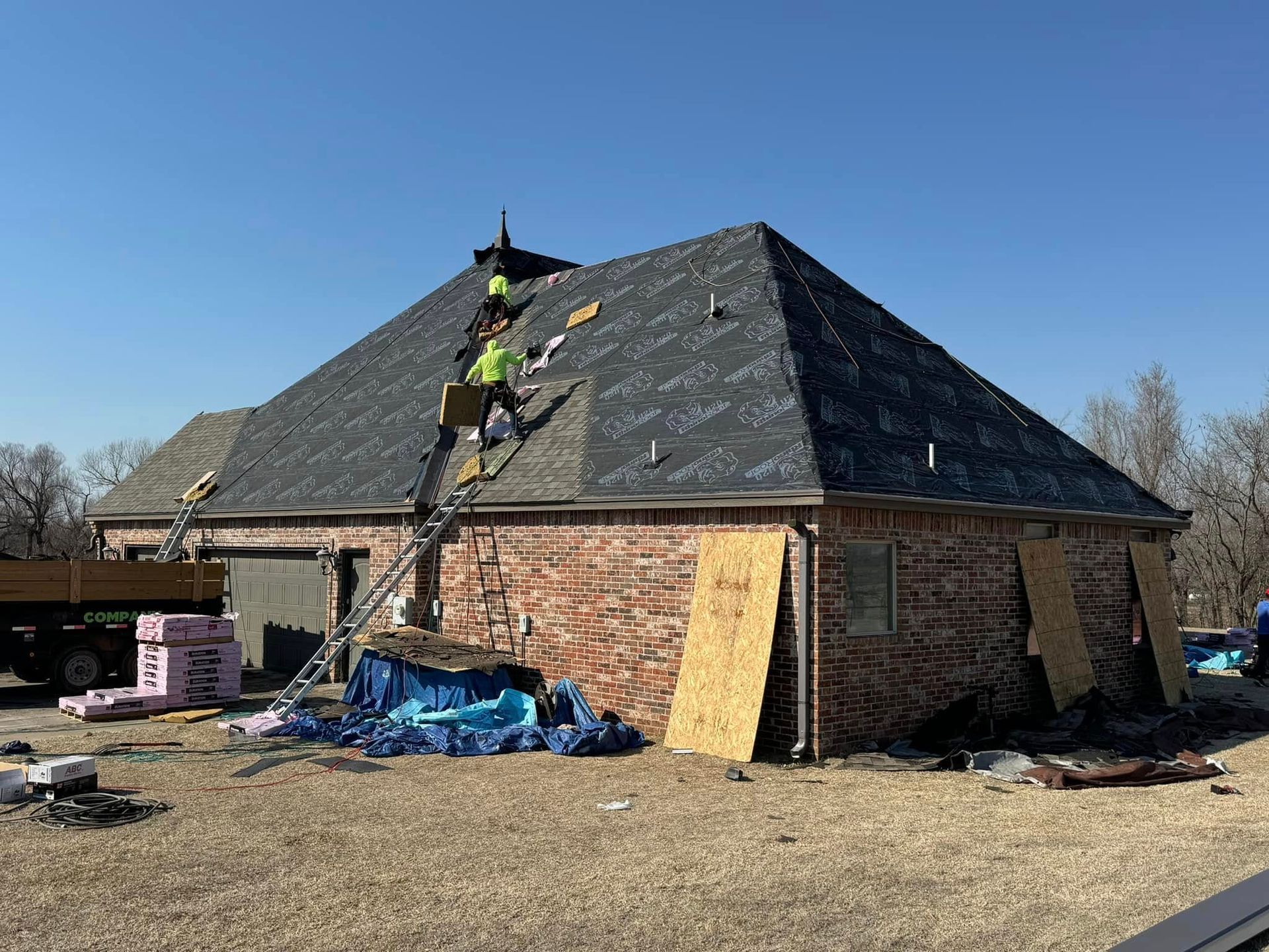 Workers in high-visibility vests install roofing shingles on a residential house under a clear blue sky.