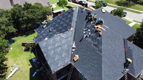 Aerial view of roofers repairing a dark-shingle house roof with materials and tools scattered around.