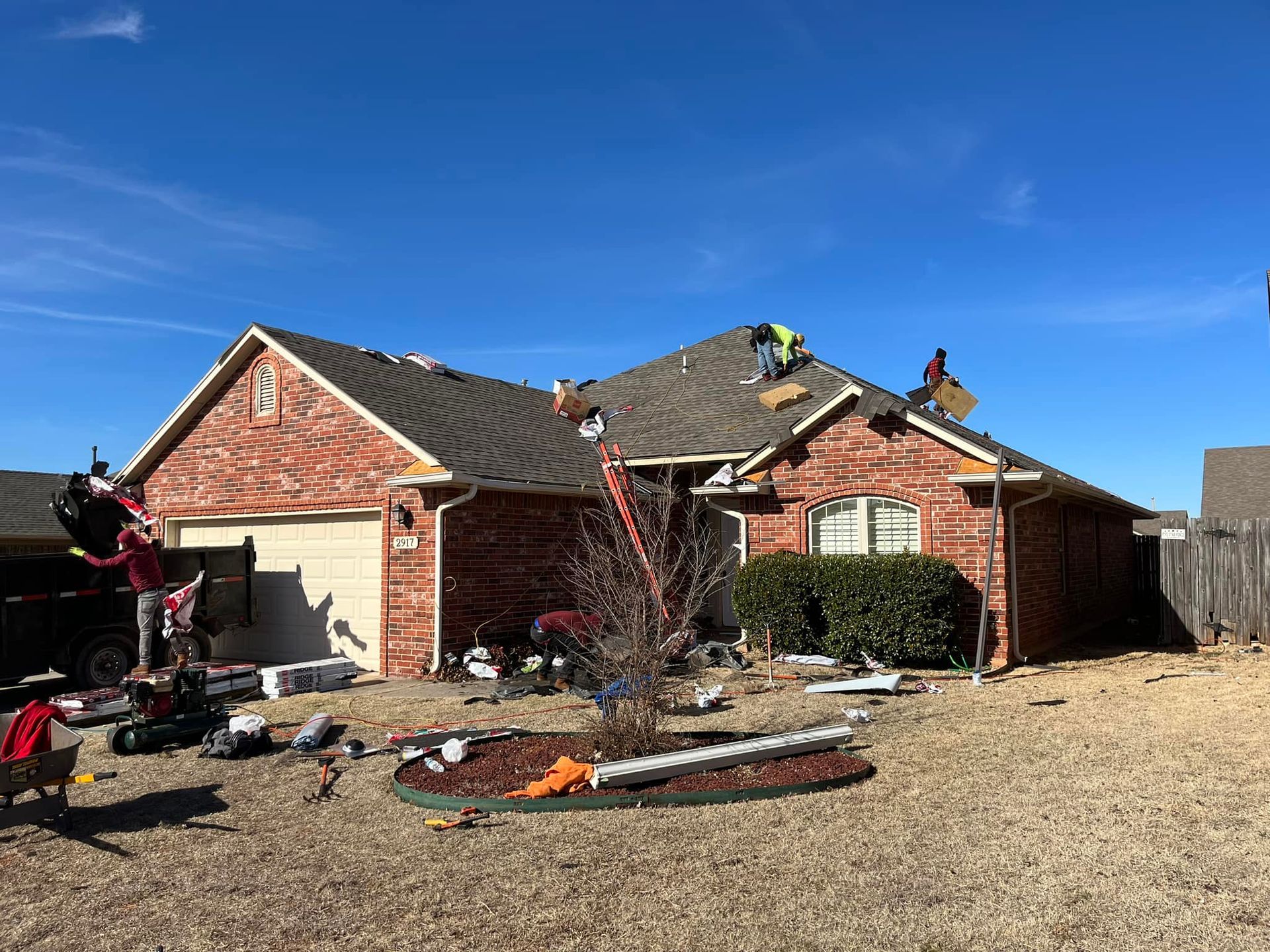 Workers replace the roof of a brick house on a sunny day, with construction materials and a ladder in the front yard.