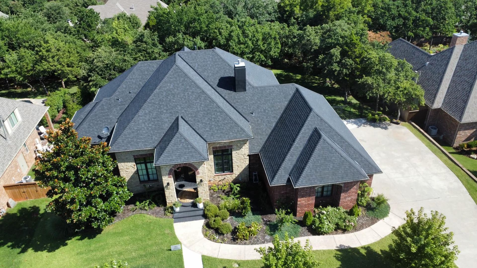 Aerial view of a residential house with a dark gray shingled roof, stone and brick siding, and a paved driveway.