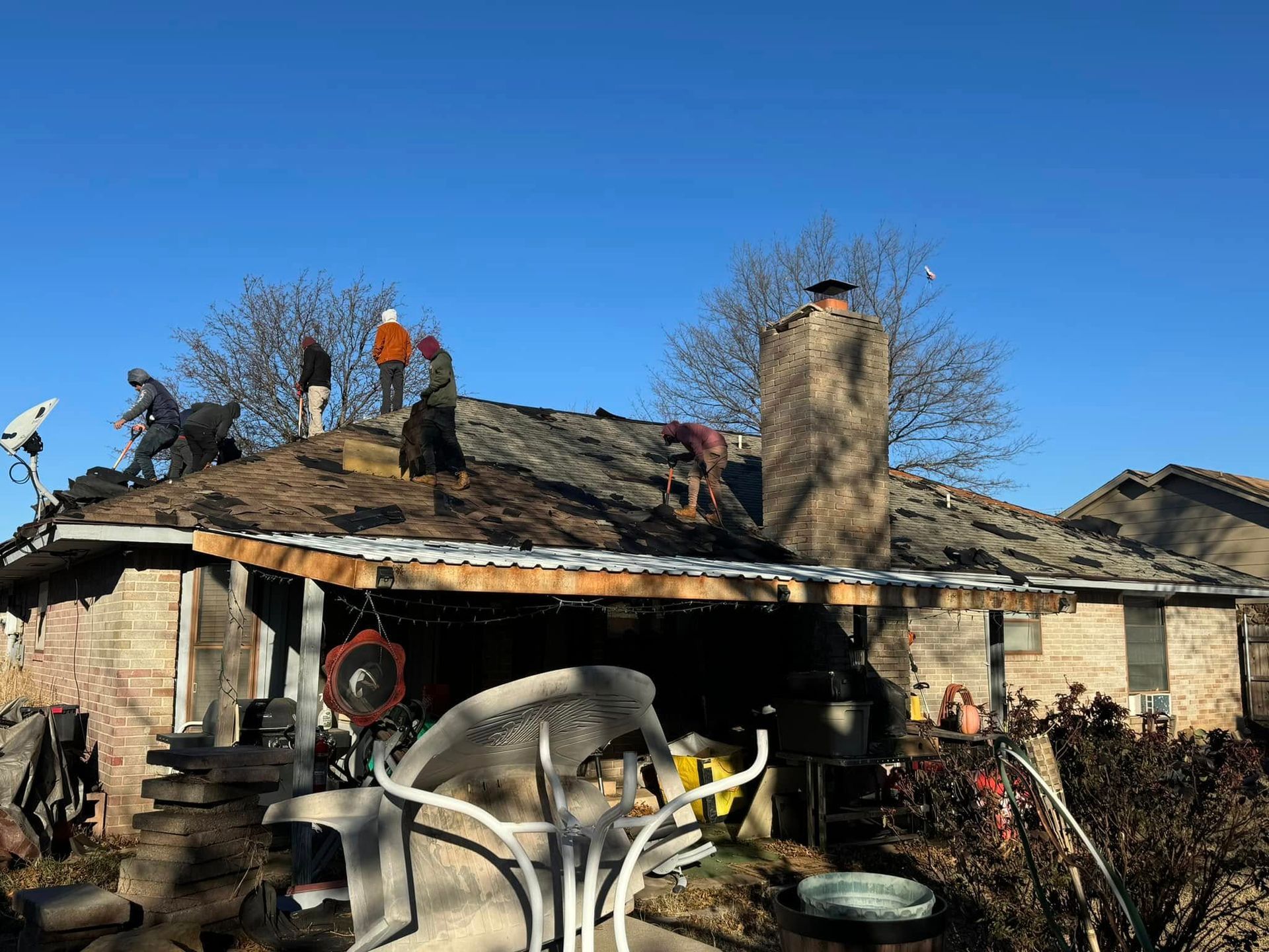 Workers in casual clothing replace shingles on a residential roof under a bright blue sky.