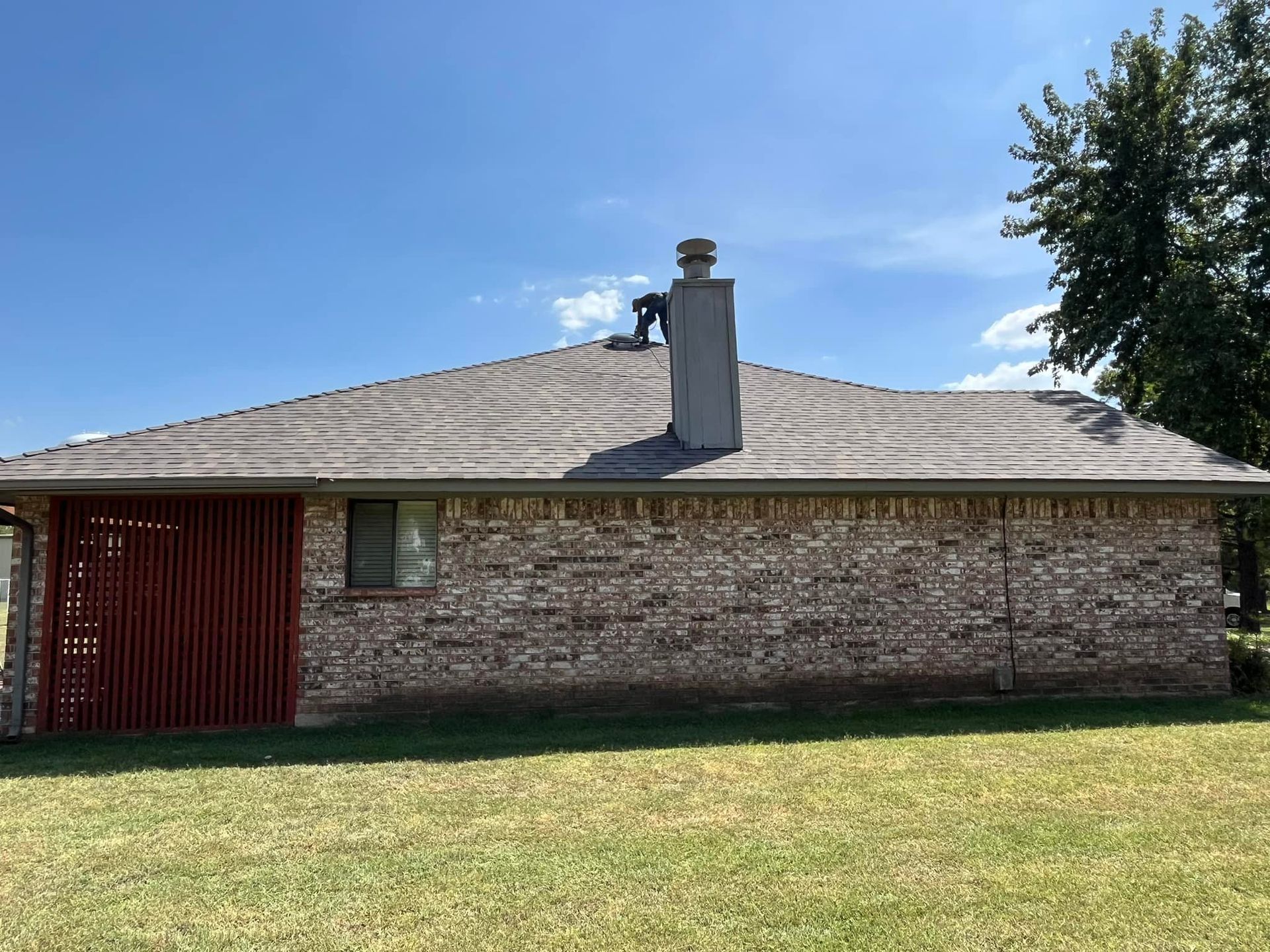 A brick house with a grey roof and a person working on the chimney under a blue sky.