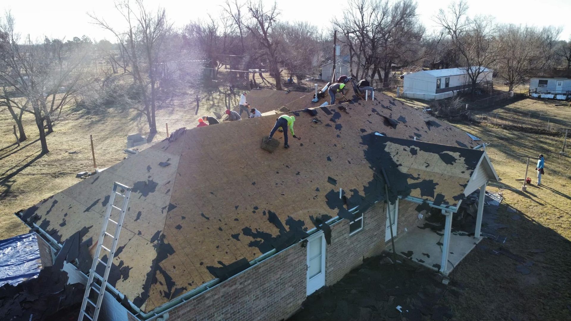 A high-angle shot of roofers removing old asphalt shingles from the wooden roof of a brick house.