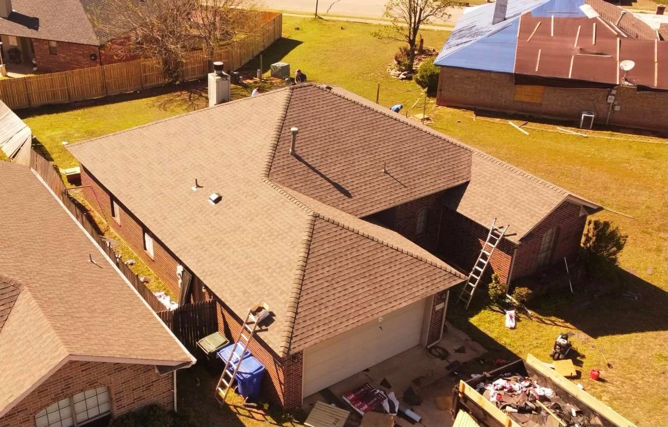 An aerial view of a brown-shingled residential roof being replaced, with a ladder and dumpster visible in the yard.