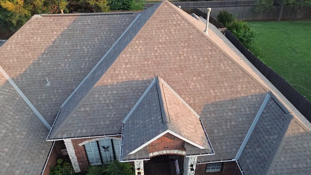 An aerial view of a brown shingled residential roof with multiple peaks and a brick front entrance below.