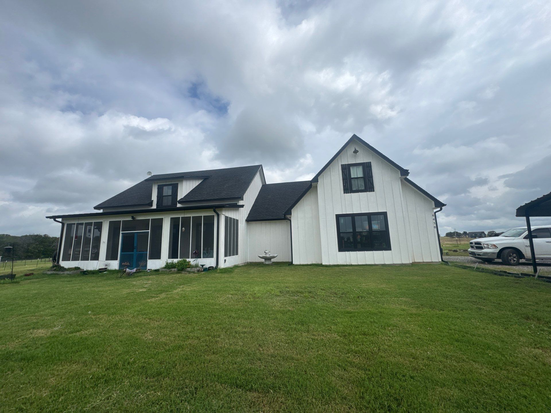 A white farmhouse with black trim and a dark roof set against a cloudy sky over a grassy lawn.