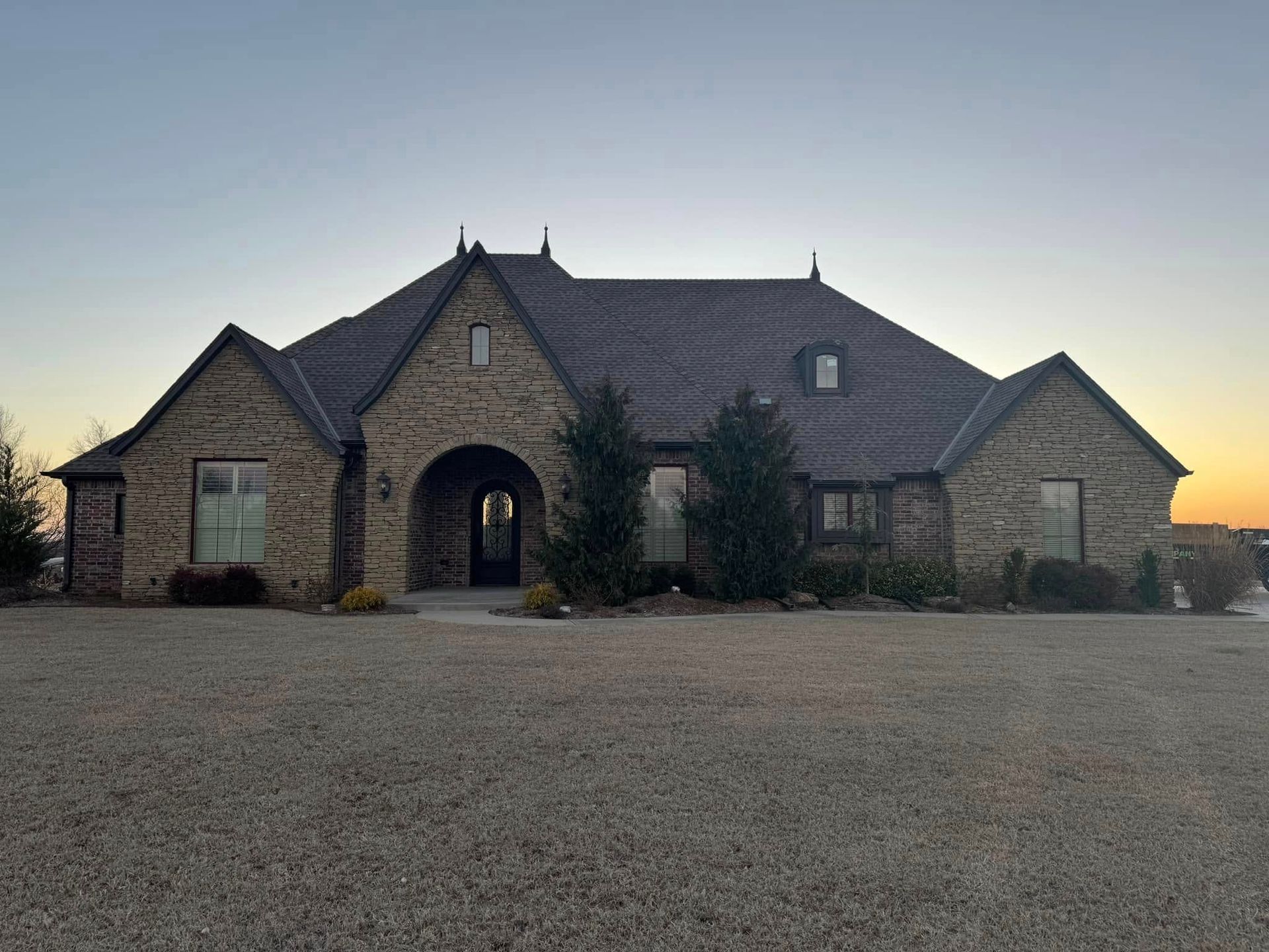 A tan stone house with a dark gabled roof and arched entryway, set against a sunset sky over a gravel lawn.