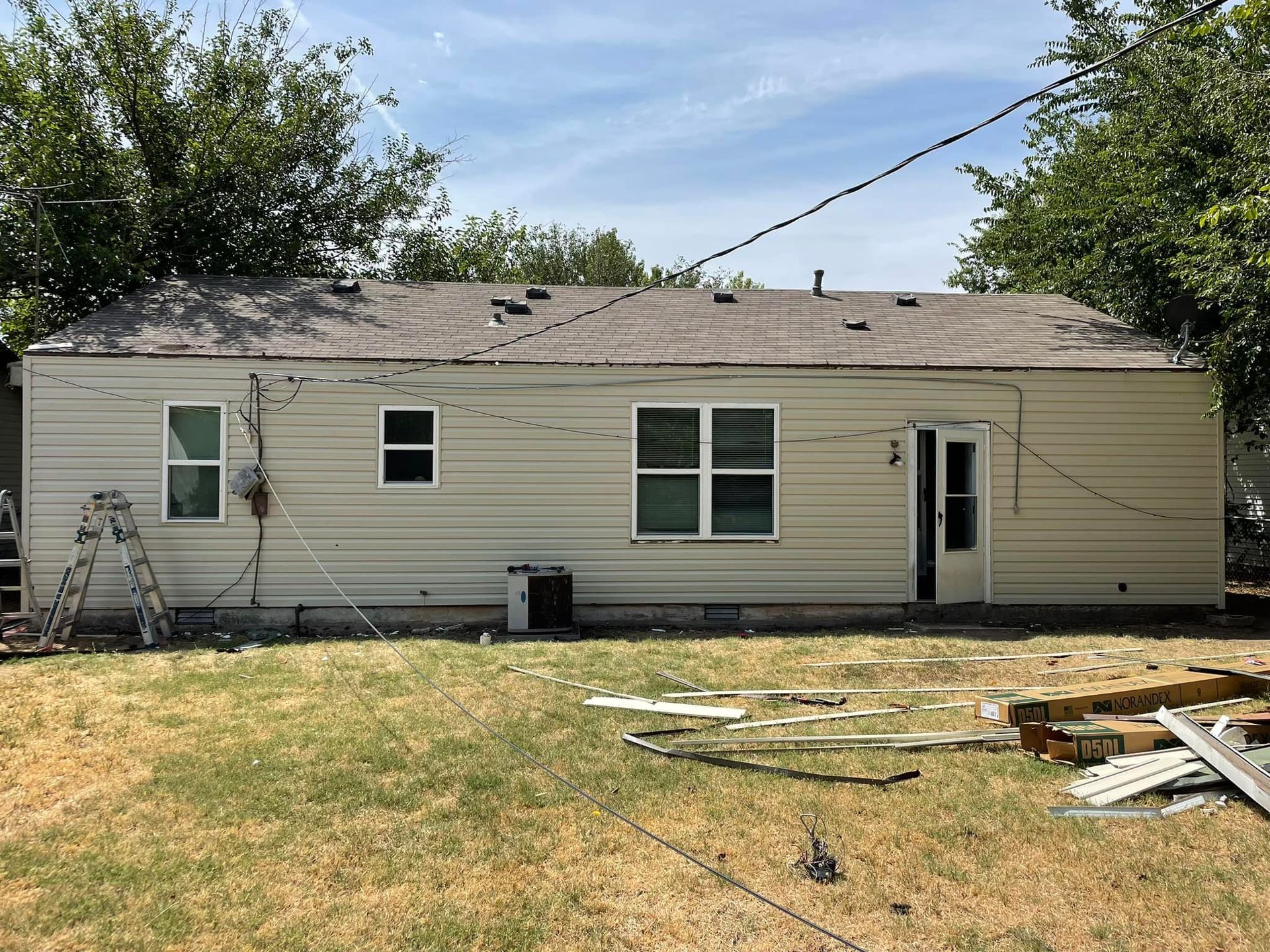 Exterior of a one-story, light-colored siding house with a weathered roof, standing in a yard with debris.