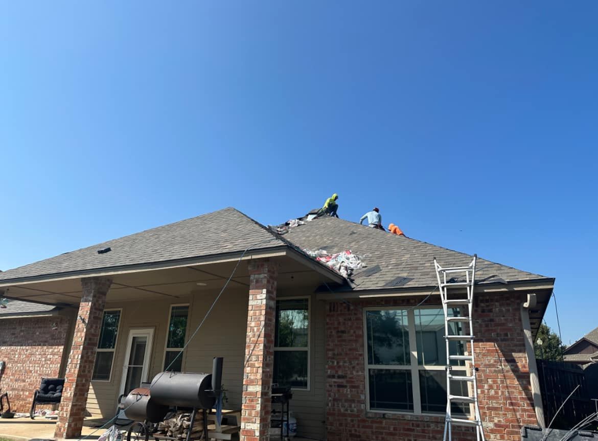 Workers repairing a shingled residential roof on a sunny day with a ladder leaning against a brick wall.