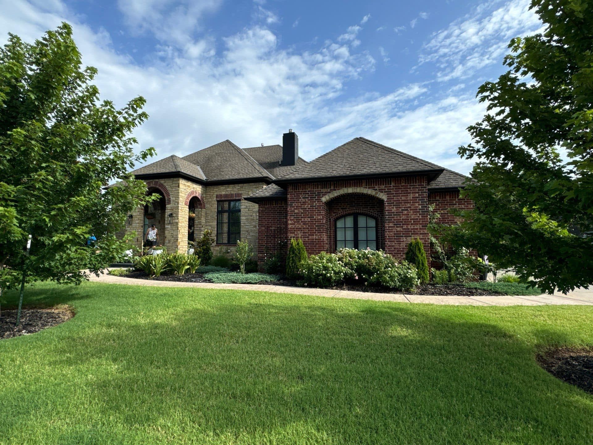 A brick and stone house with a dark shingled roof, set behind a lush green lawn on a sunny day.