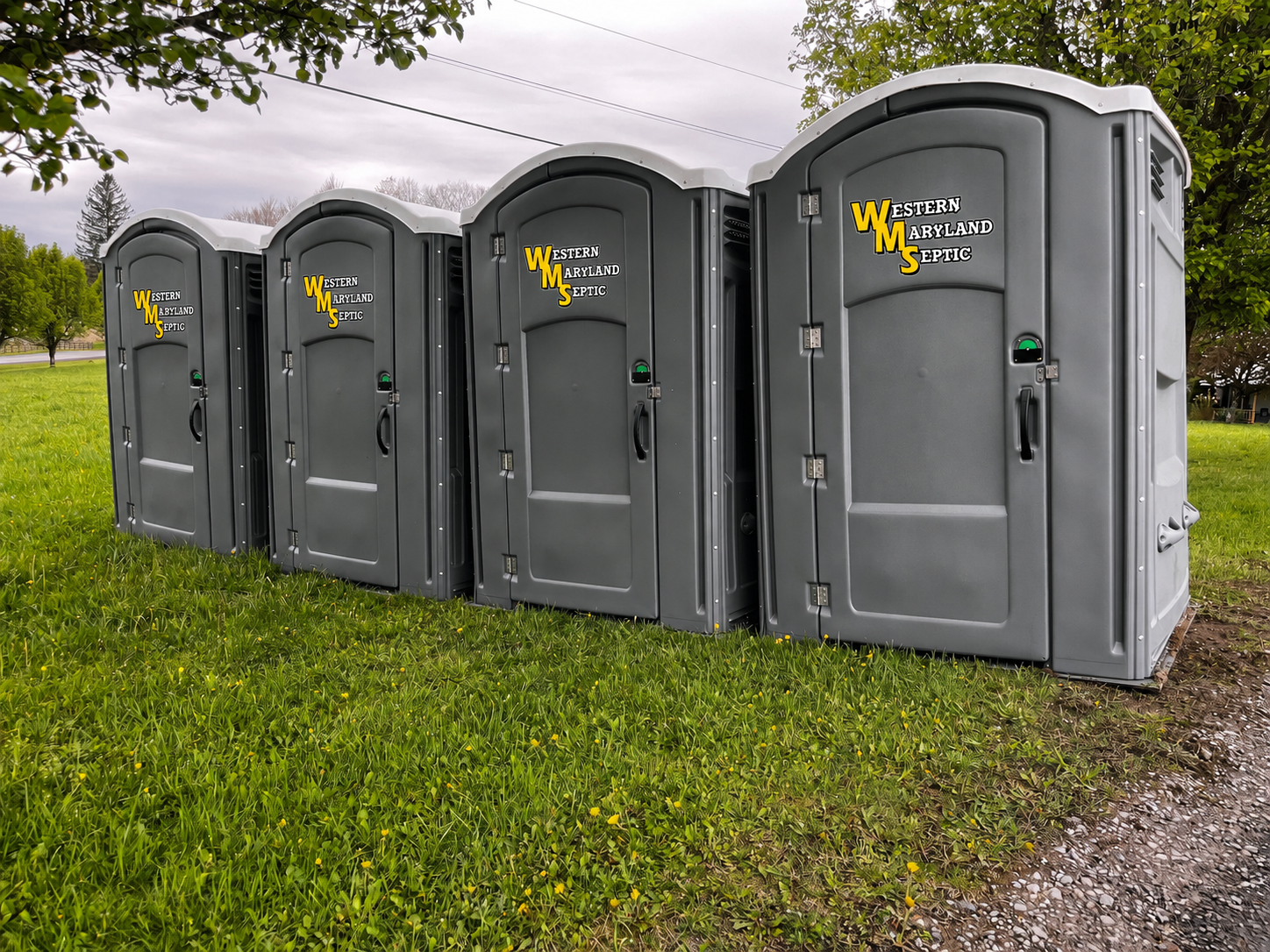 Four gray portable toilets lined up on grass outdoors under a cloudy sky.