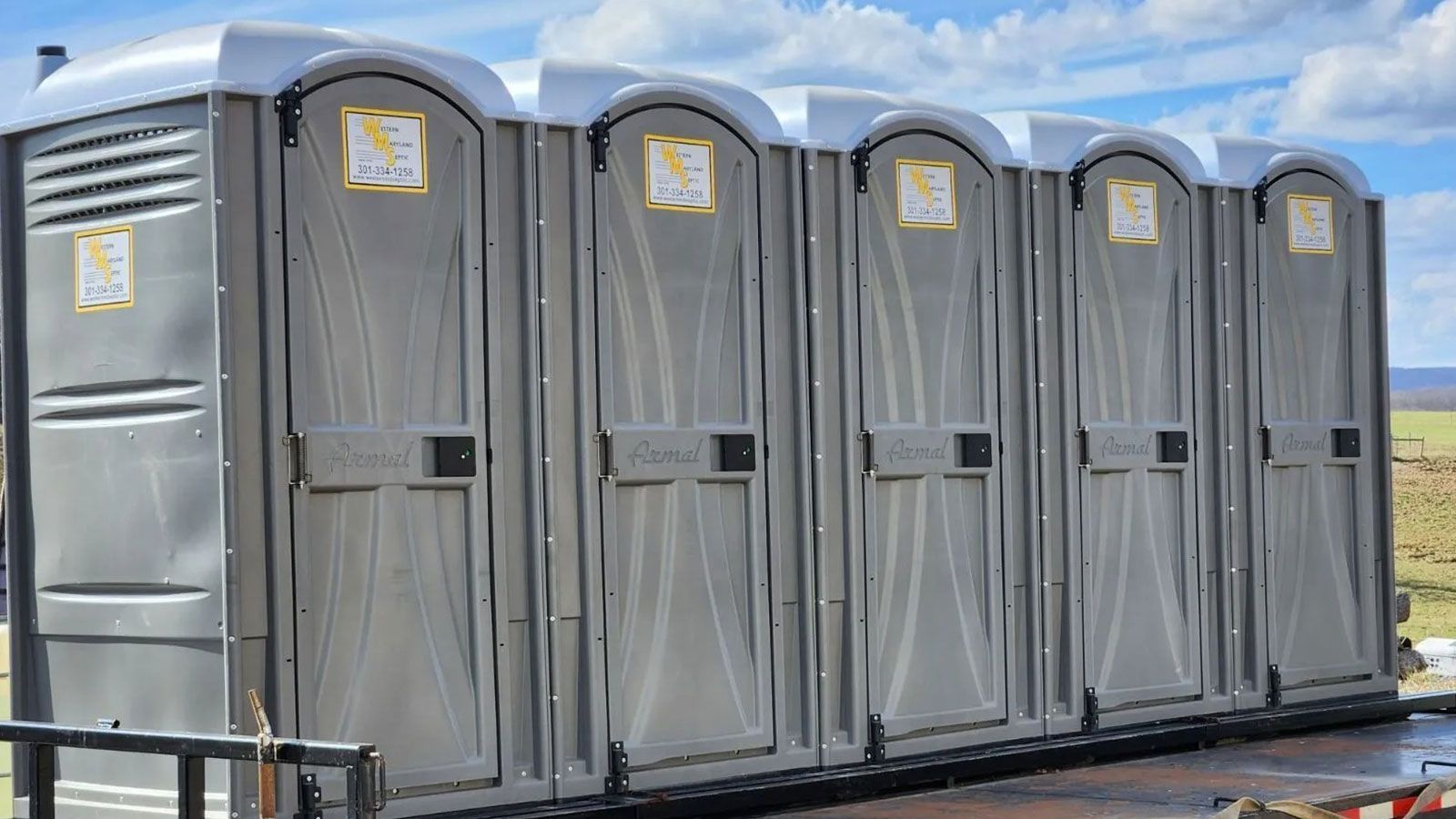 Row of gray portable toilets on a trailer, parked outdoors against a blue sky