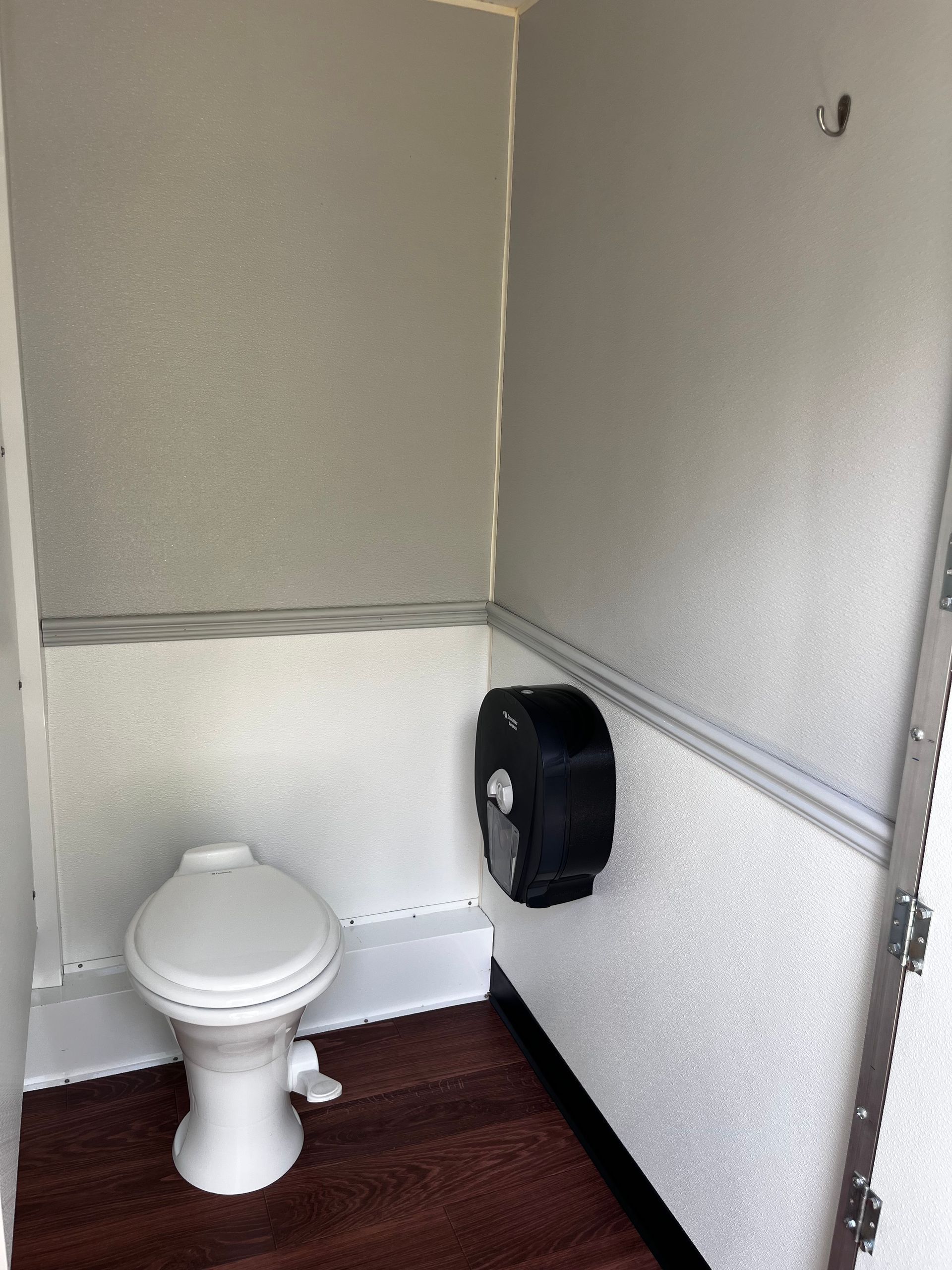 Small restroom with a white toilet, black toilet paper dispenser, and dark wood-patterned floor.
