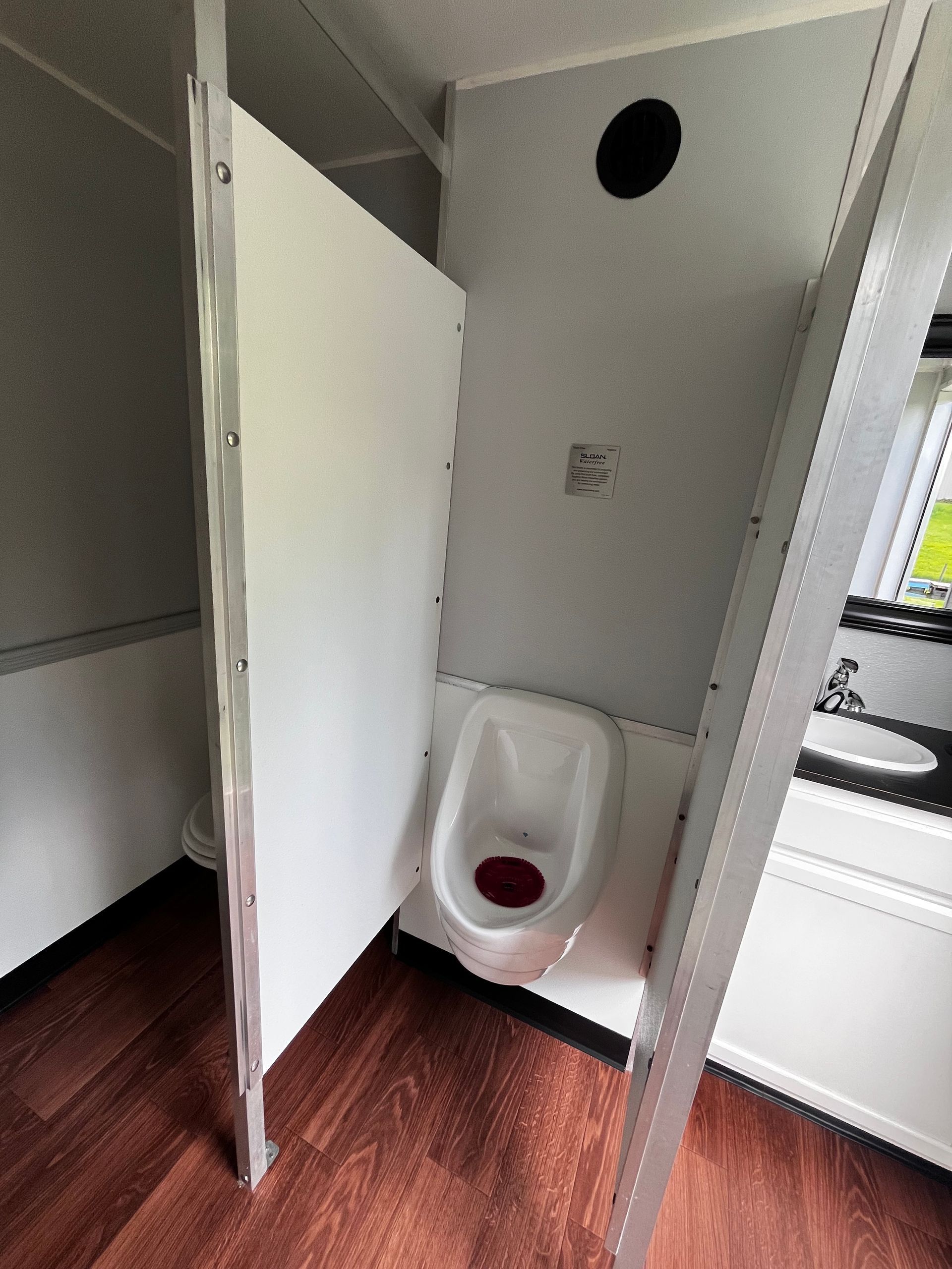 Public restroom stall with open white partition and toilet with red seat insert on wood floor.