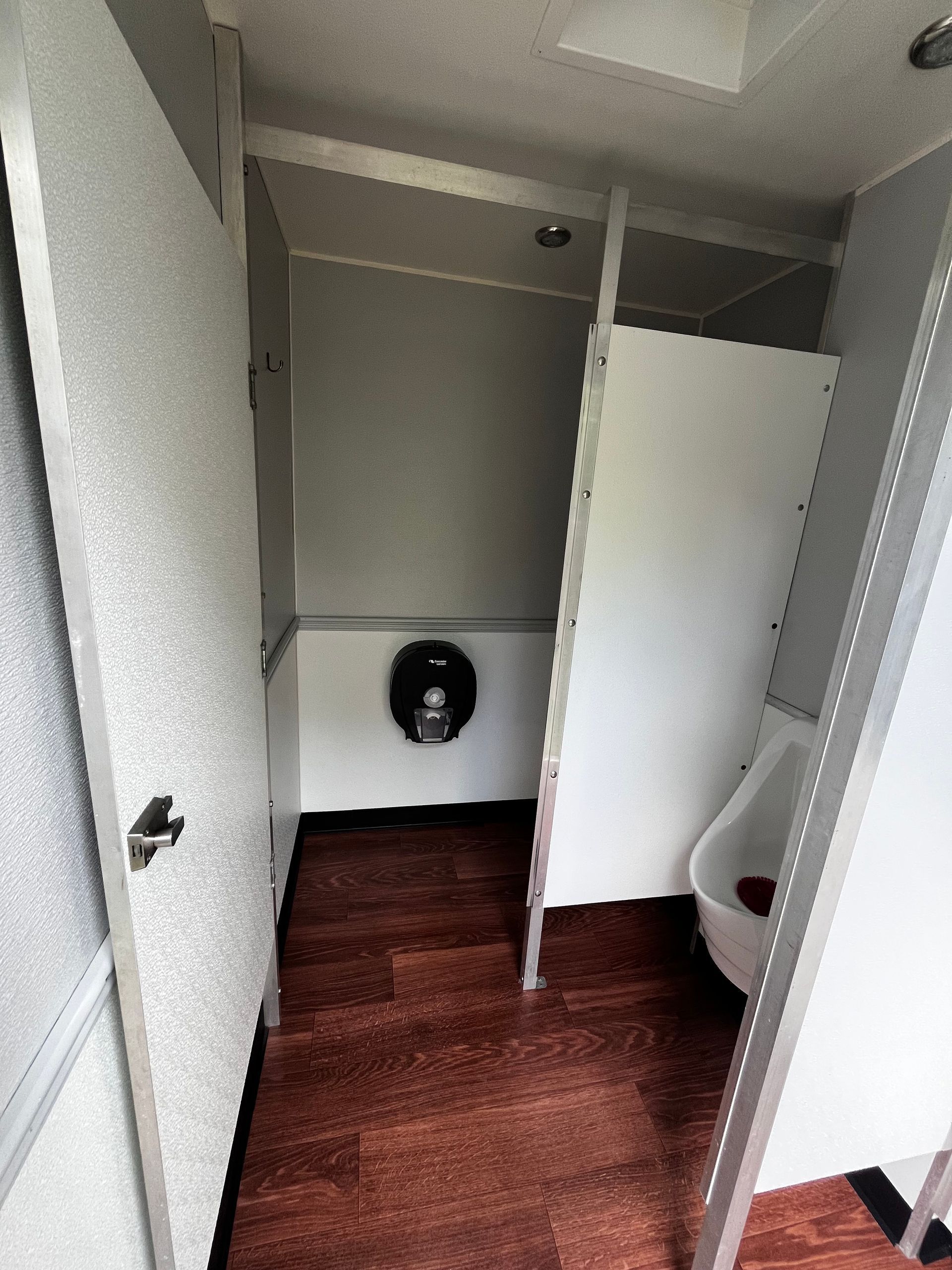 Small restroom with open stall door, white walls, dark wood floor, and wall-mounted toilet paper holder.