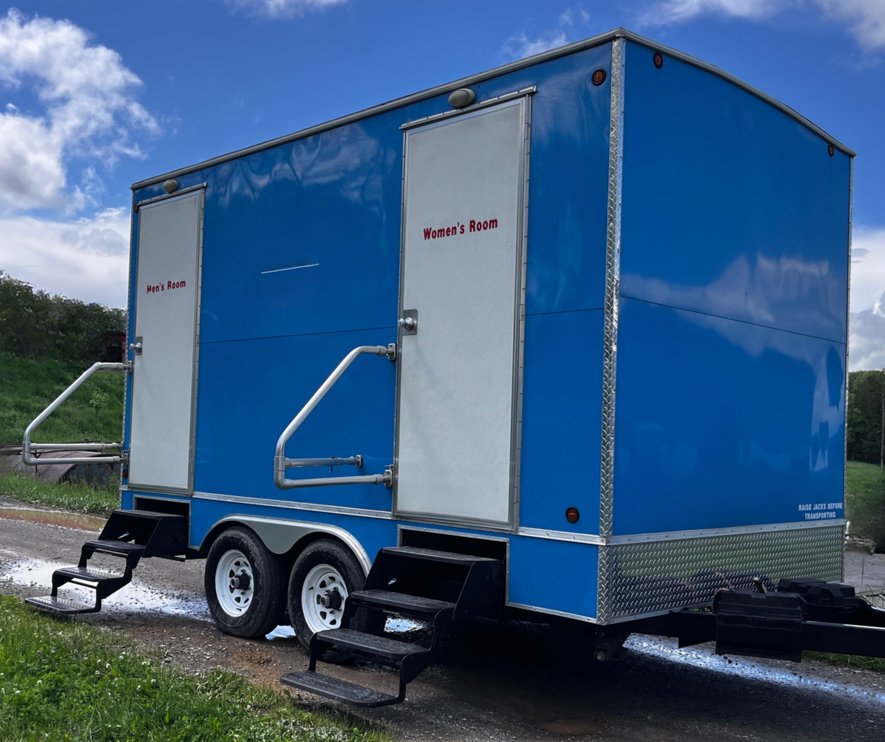 Blue portable restroom trailer with silver doors and steps, parked on a grassy roadside under a cloudy sky