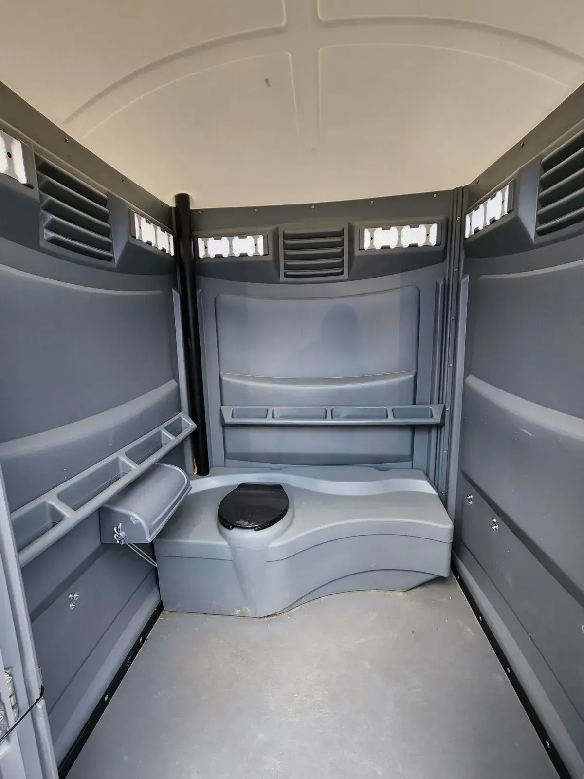 Interior of a grey portable toilet, with a toilet, shelves, and vents.