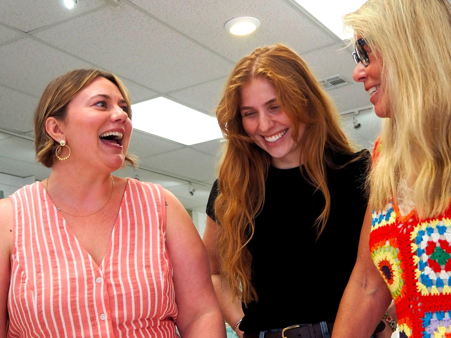 Three women smiling and laughing together indoors under bright ceiling lights.